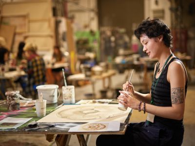 In the foreground towards the right, a person with short brown hair wearing a striped top and a tattoo on their arm sits at a desk with a paintbrush in one hand and a cup of water in the other. On the desk is a painting they are working on, as well as other paints and designs to copy from. In the background you can see other individuals at desks blurred in a studio.